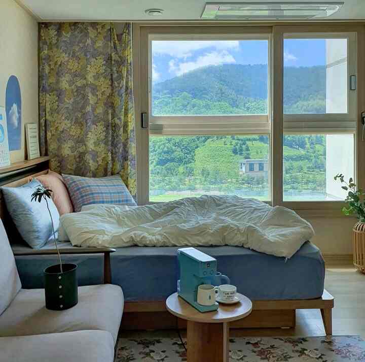 Natural-toned bedroom with wooden furniture and white bedding, featuring large window overlooking lush nature view