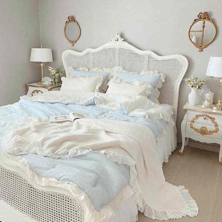 ホワイトとゴールドのアクセントが映えるフレンチカントリースタイルの寝室、ラッフル付きのブルーの寝具と枕が配置された温かみのあるインテリア