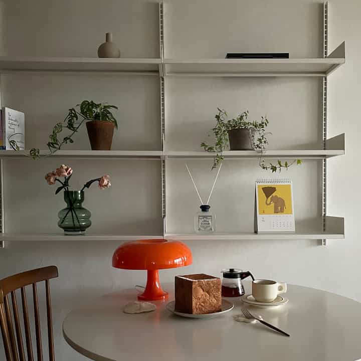 White-toned dining room featuring a round table, wooden chair, and shelves with plants and decor, creating a simple natural vibe