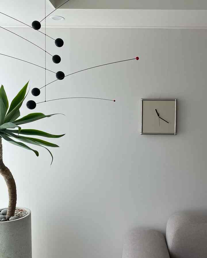 白い壁と天井のミニマルなリビングルーム、左に大きな緑の植物、右に壁時計とグレーのソファが配置された印象的な空間