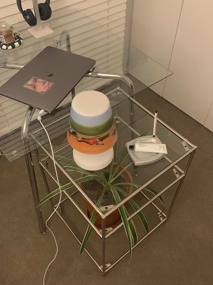 Modern indoor space with transparent glass table featuring a plant on a modular shelf, brown carpeted floor and white blinds