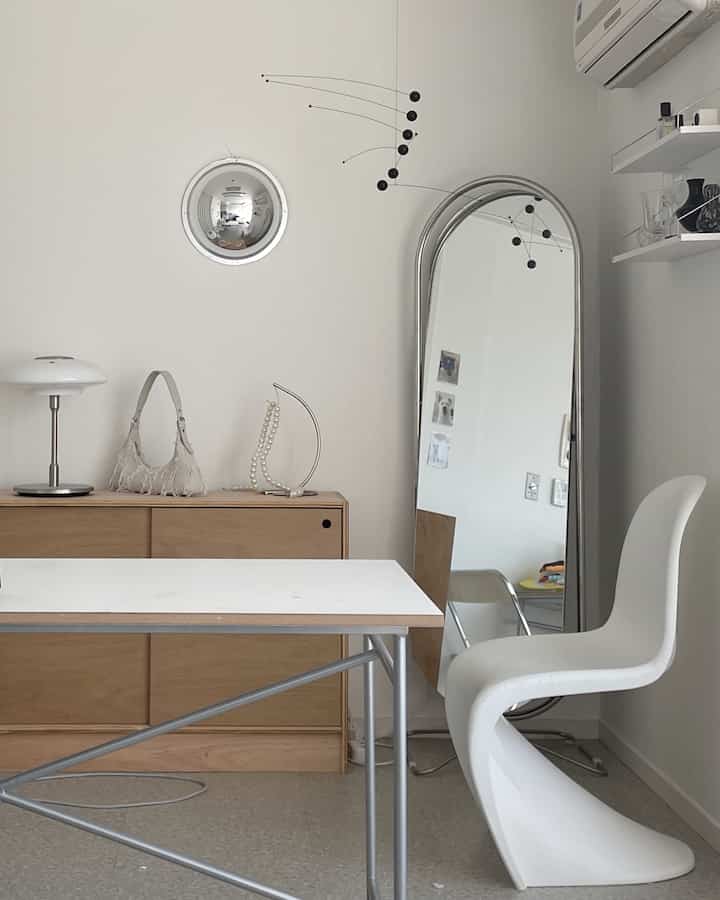 White and wood-tone studio apartment featuring a simple desk, dresser, and full-length mirror with a clean and minimalistic interior
