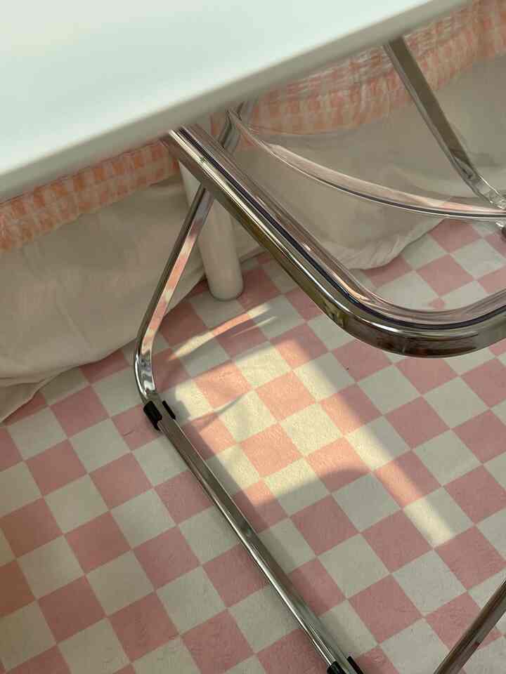 A studio apartment space with a pink and white checkered rug, featuring a transparent folding chair in a bright and cozy atmosphere