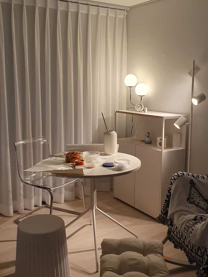 White-toned living room featuring round table, transparent chair, sofa, and warm lighting creating cozy home cafe atmosphere