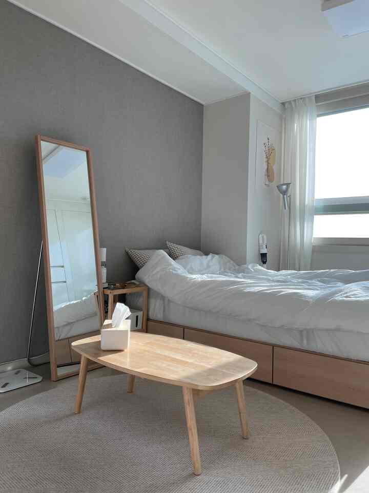 Natural wood tones and white dominate this small studio bedroom featuring simple furniture and a large window, creating an airy space