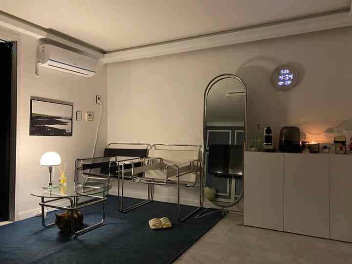 Studio apartment living room with white walls and a blue rug, featuring metal frame armchairs and a glass table creating a cozy atmosphere