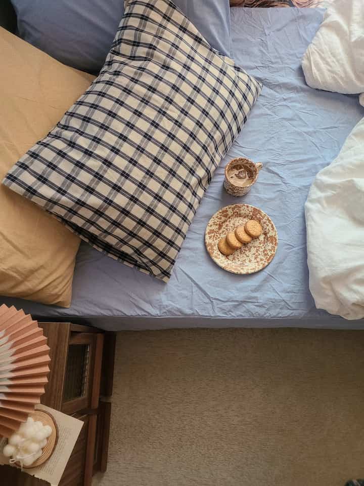 A cozy bedroom with light blue bed sheets, beige carpet, and checkered pillows, featuring marble-patterned mug and plate in a home cafe setup