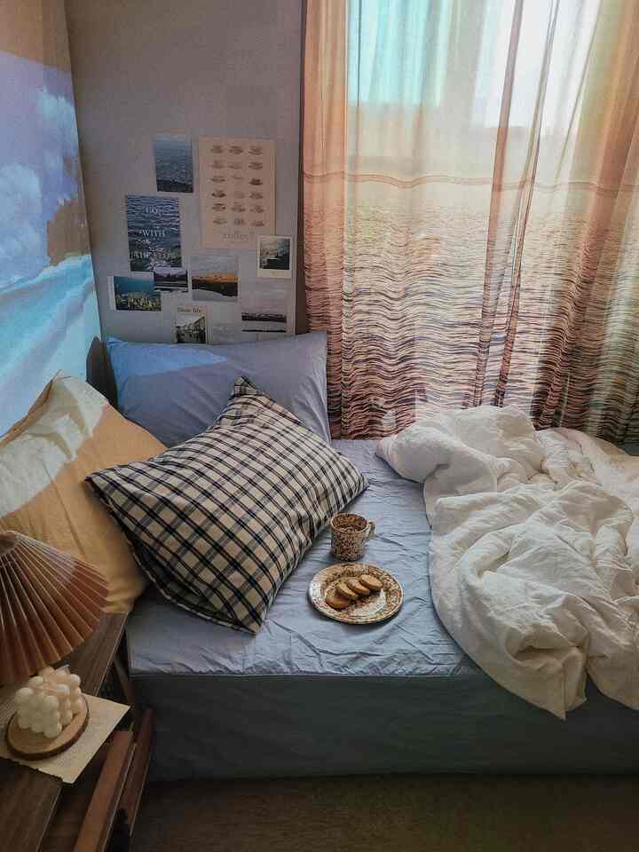 Studio bedroom with sunset light filtering through patterned curtains, featuring blue bedding and wall posters in cozy setting