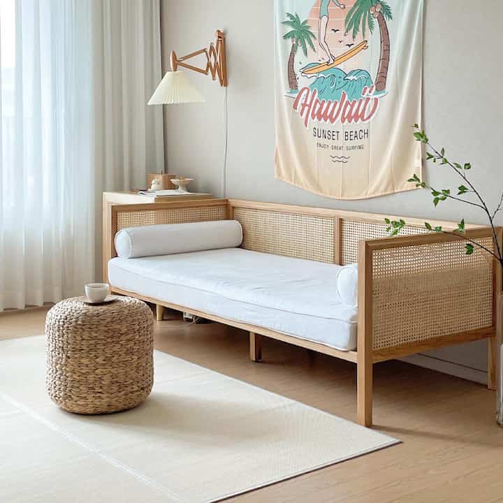 ベージュと木目調のナチュラルなワンルームリビング、ラタンソファとスツール、夏を感じさせるファブリックポスターが配置された雰囲気のあるインテリア