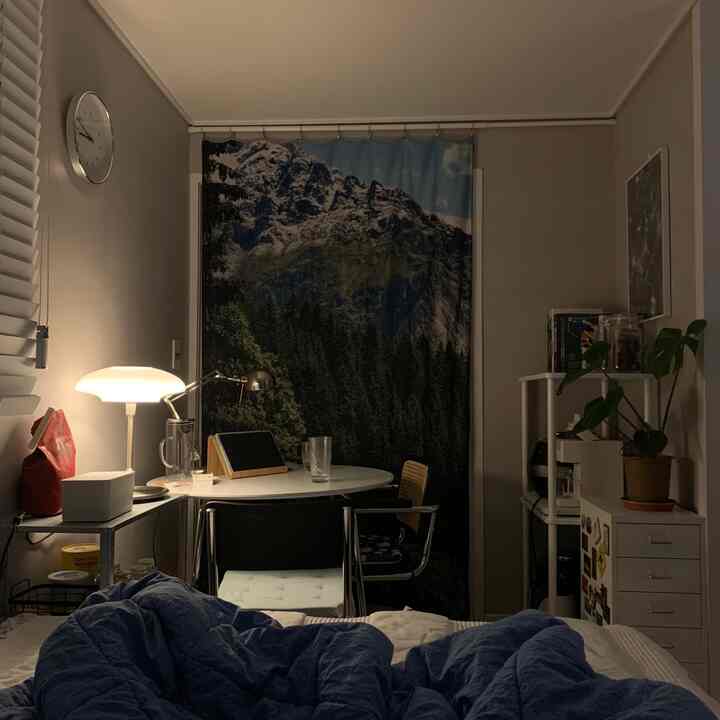 柔らかなベージュの小さな寝室、山の風景カーテンとホワイトデスク、暖かい光のテーブルランプが配置された雰囲気のあるインテリア