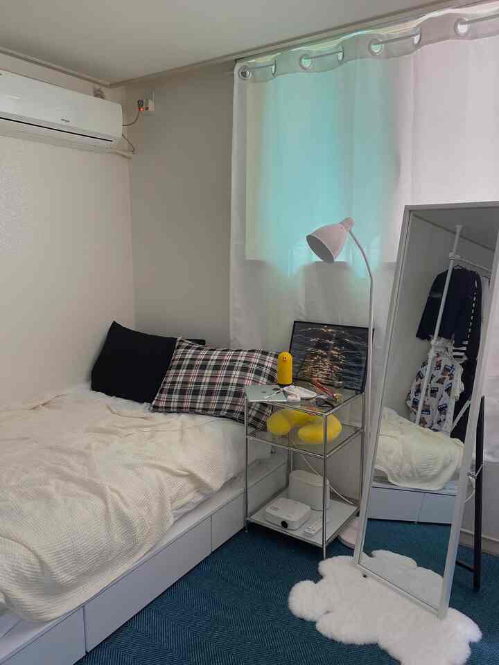 Compact single bedroom in white and navy tones featuring a bed, side table, full-length mirror, and floor lamp with a cozy atmosphere