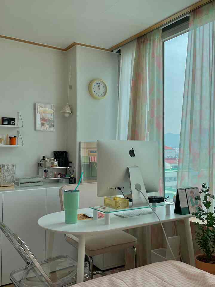 A studio apartment home office in white and pastel tones featuring a large window with curtains and an iMac computer on a clean white desk