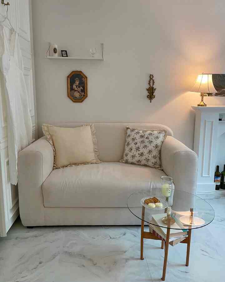 Beige-toned living room featuring a beige sofa and glass-top side table, wall art, and warm lamp lighting creating cozy ambiance