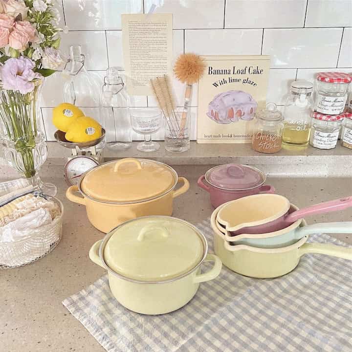 Pastel yellow, green, and pink enamel pots and kitchen utensils arranged on a kitchen counter, creating a cozy home cafe vibe