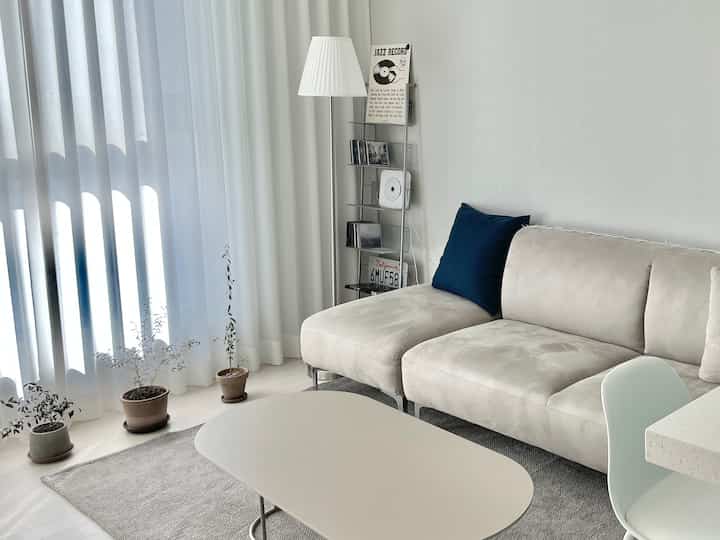 A white and gray toned living room featuring a beige sofa, coffee table, curtains, and potted plants by the window creating a cozy atmosphere