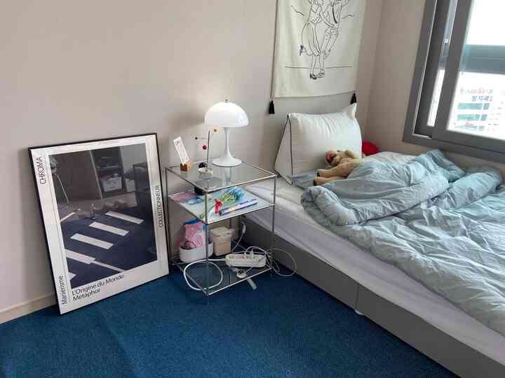 A blue-carpeted studio apartment with white walls, featuring a metal frame side table next to a bed with bedding and a dog plush, creating a cozy bedroom