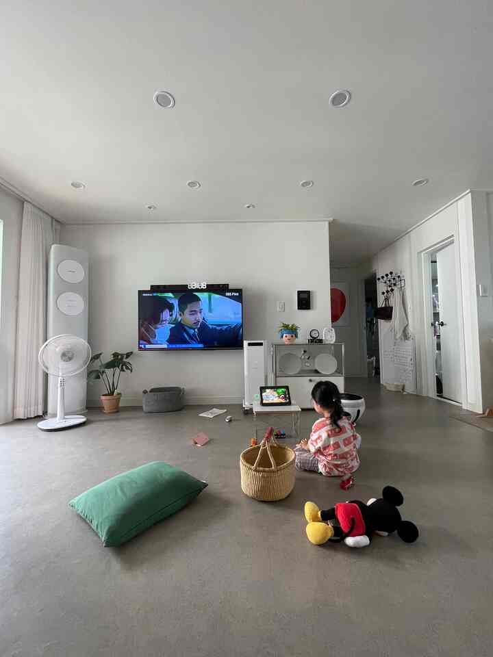 White-toned living room featuring a child sitting on the floor watching a video amidst toys and cushions in a slightly cluttered space