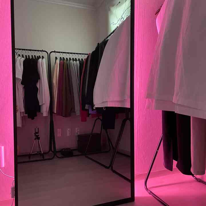 ピンクのネオンライトが柔らかく照らすウォークインクローゼット、黒のハンガーと白い服が整然と並ぶモダンな空間