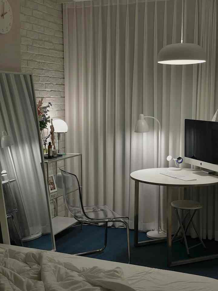 White-toned studio bedroom and home office space featuring a transparent chair, round table, and pendant lighting in a simple modern interior
