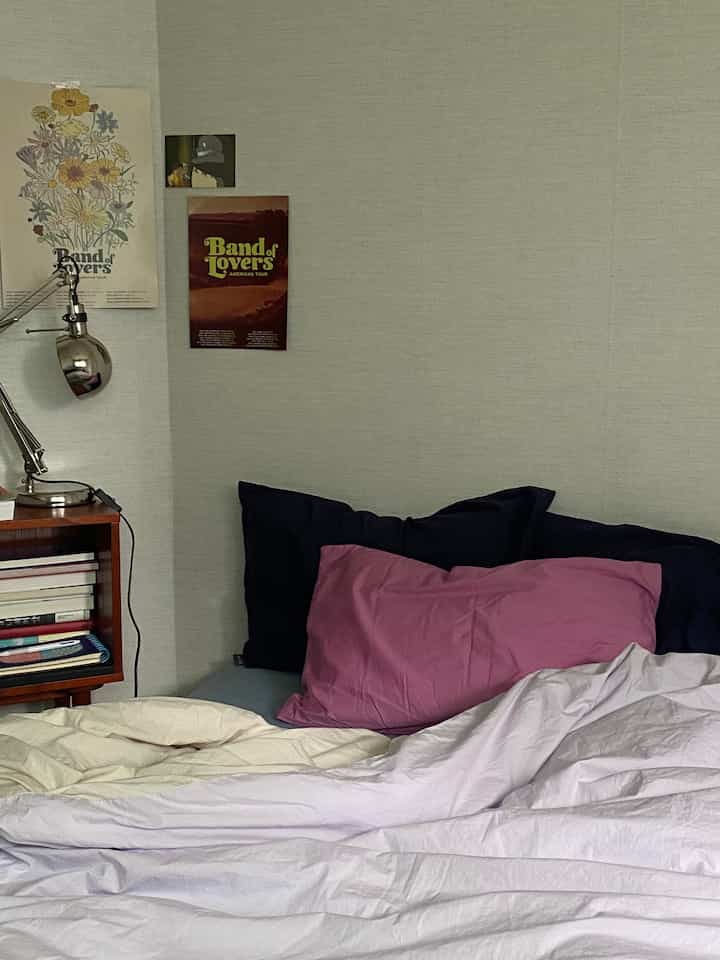 柔らかなベージュと白のトーンの寝室、ピンクとネイビーの枕が並ぶベッドとレトロなポスターが配置された雰囲気のある空間