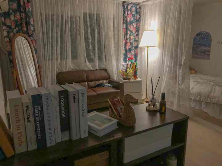 Vintage-style living and bedroom combined space featuring a brown sofa, floral lace curtains, and a floor lamp with cozy atmosphere
