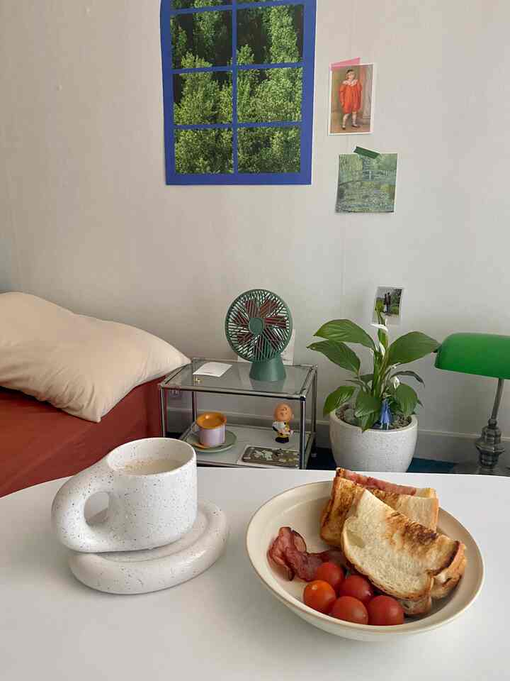 白い壁と緑のアクセントが調和する居心地の良いリビング、ソファベッド、卓上扇風機、マグカップと皿が置かれたテーブルがある雰囲気のあるインテリア