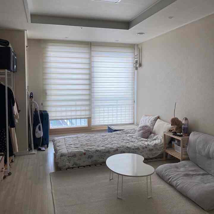 ホワイトとベージュのトーンの小さな寝室、ベッドとソファ、丸テーブル、犬のぬいぐるみが置かれたシンプルな空間