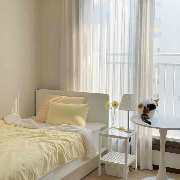 White and yellow toned bedroom featuring a bed, round table with a cat by a window with sheer curtains