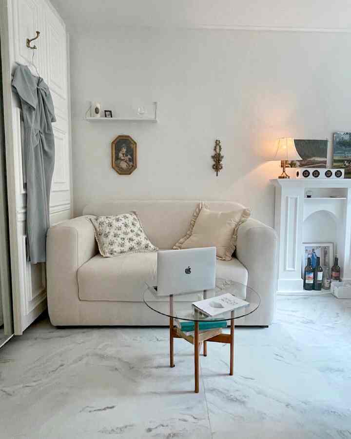 明るいアイボリー調のリビング、クリーム色の2人掛けソファと円形ガラステーブル、ナイトテーブルと額縁が配されたシンプルモダンなインテリア