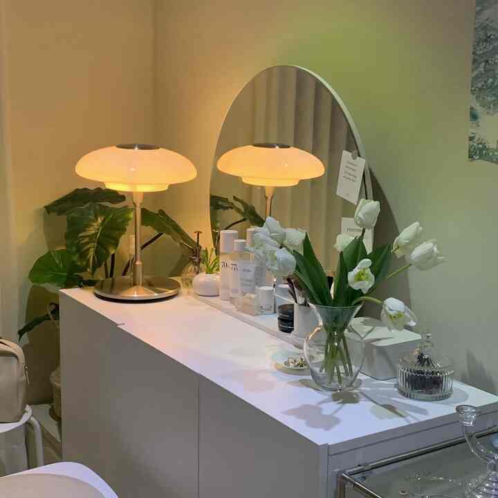 White and green bedroom vanity featuring table lamps, an oval mirror, plants, and a clear flower vase for a cozy atmosphere