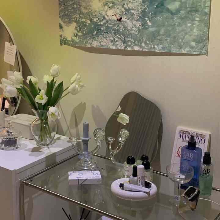 White and transparent glass-toned powder room featuring mirrors, a vase with flowers, and candles, creating a neat and calm atmosphere