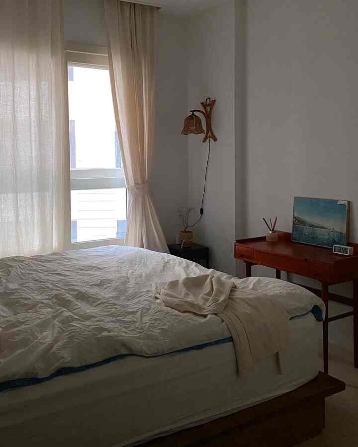 Beige and white toned bedroom with large window featuring linen curtains and wooden console table, creating a natural relaxation space