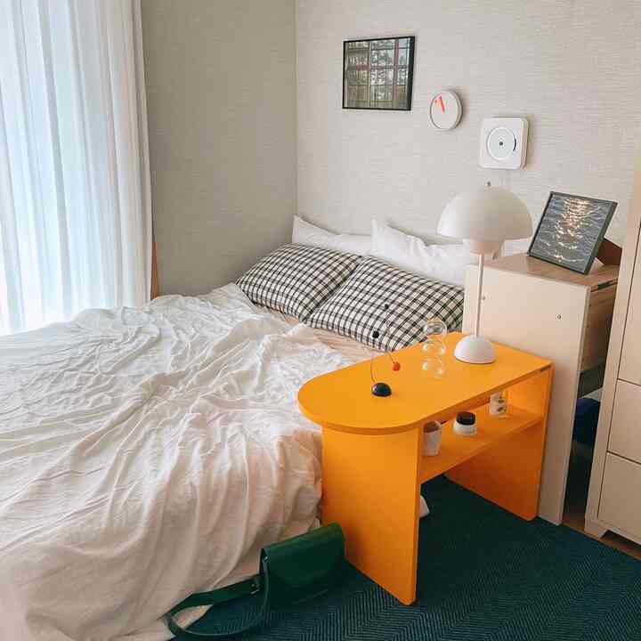 ホワイトの寝具とカーテンがある寝室、黄色のサイドテーブルとシンプルなモダンランプが配置されたコンパクトな空間