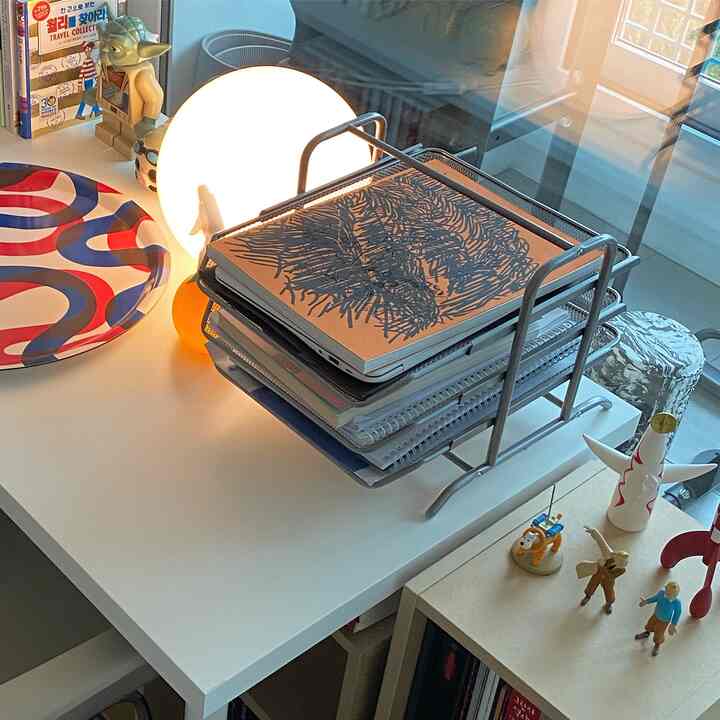 White-toned desk space featuring books, a multi-tier tray, and a round table lamp in a tidy home office setting