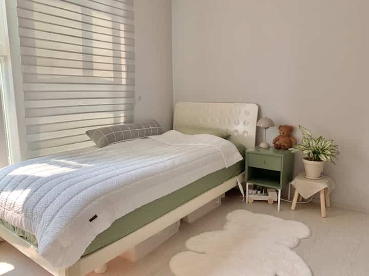 A cozy bedroom in white and green tones featuring a bed, nightstand with table lamp, plant, and teddy bear in a kids' room setting