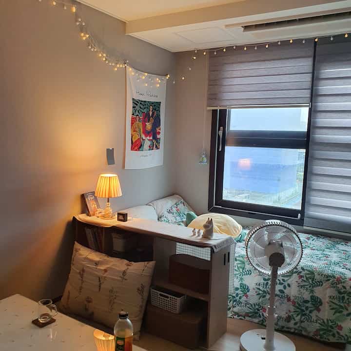 暖かいベージュとグレーのトーンのワンルーム寝室、居心地の良い照明と本棚の仕切り、扇風機が配置された雰囲気のあるインテリア