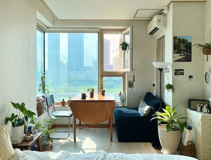 ナチュラルトーンのコンパクトなリビングルーム、窓際に木製のダイニングテーブルとダークカラーの一人掛けソファが配置された落ち着いた空間