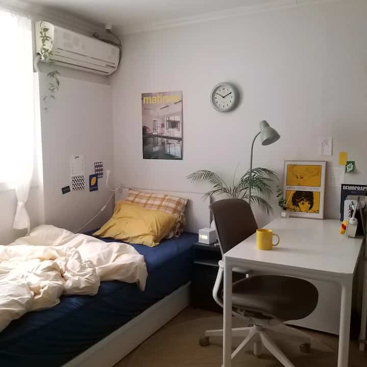 A cozy studio apartment bedroom featuring a simple modern desk and bed in soft bright tones with navy accents