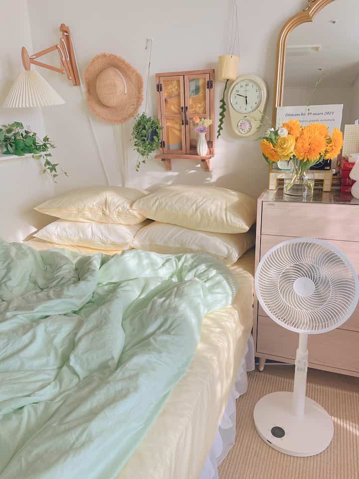 Natural tone bedroom featuring cream and mint bedding, wall-mounted clock, plants, and a standing fan creating a cozy atmosphere