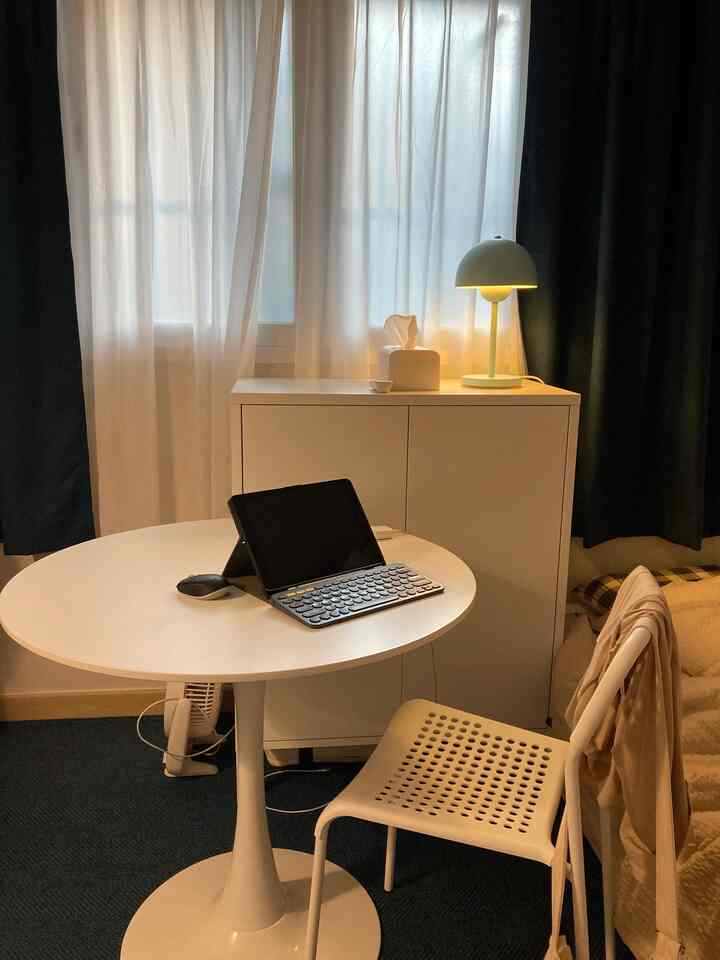 White-toned studio apartment living space with round table, chair, cabinet, and lamp arranged in a simple setting
