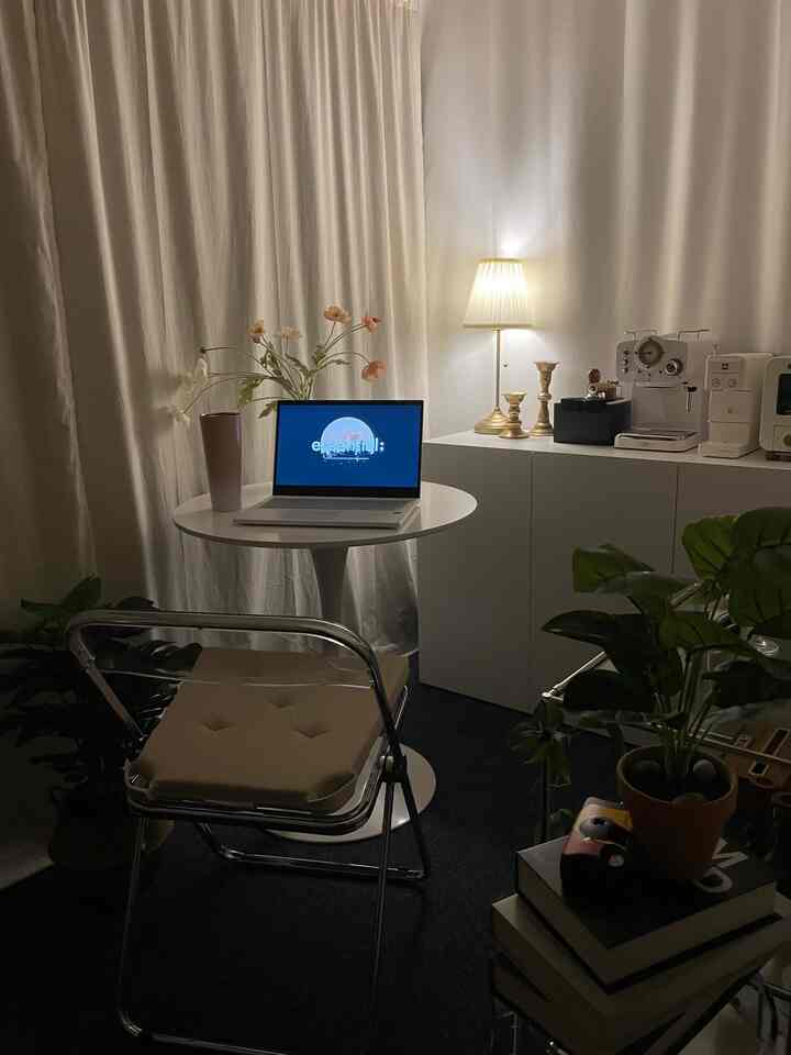 柔らかなベージュとホワイトのワンルーム空間、透明な椅子と丸テーブル、温かみのある照明とインテリア小物が配された居心地の良いおうちカフェのインテリア