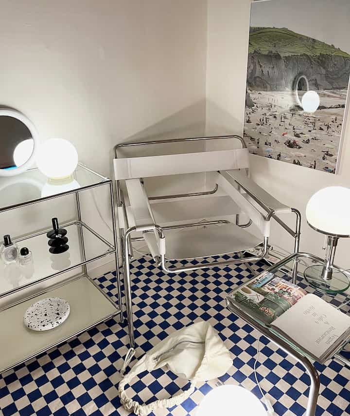 白とクロム家具が印象的なワンルーム、チェッカーボード柄のラグとポスターが調和するモダンなインテリア