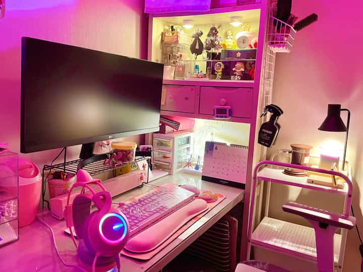 ピンクの照明で満たされた狭いデスク空間に、コンピューター、ヘッドセット、多様なかわいい装飾が置かれた雰囲気のあるゲームルームのインテリア
