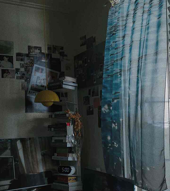 Small bedroom-like space with blue-tone curtains and a yellow pendant light, featuring posters and book stacks adding cozy vibes