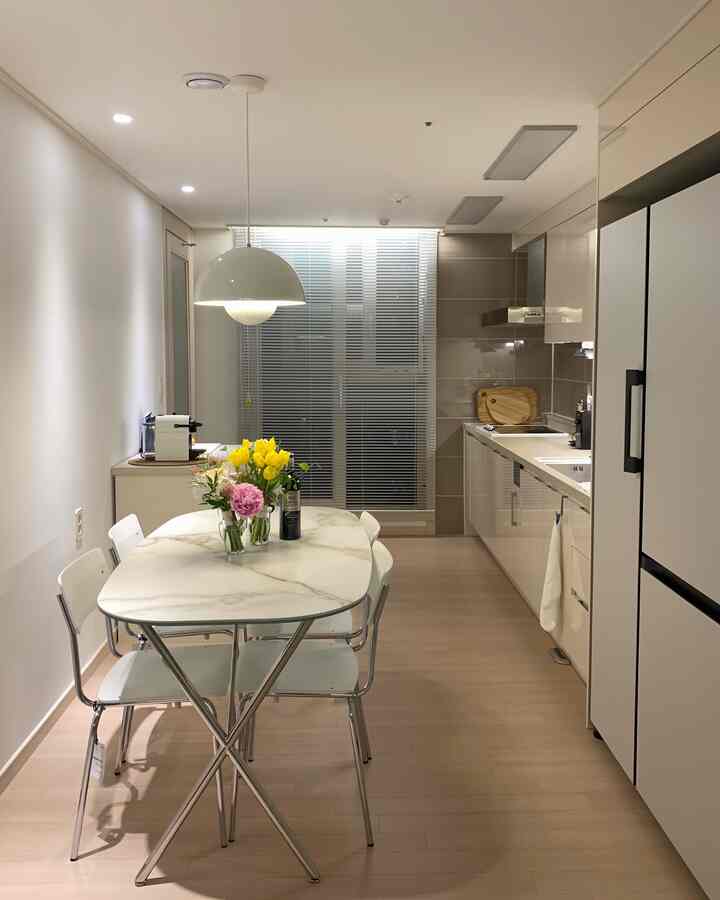 A clean, modern kitchen and dining room in white and wood tones, featuring a marble dining table and silver aluminum blinds