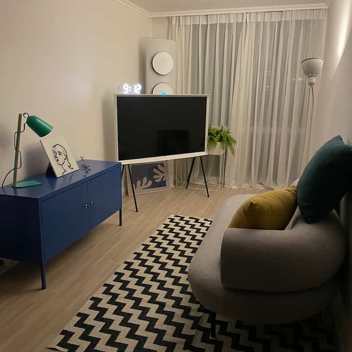 Bright white walls and wood-tone floor define a compact living room featuring a blue cabinet, gray two-seater sofa, floor lamp, and patterned rug with cozy atmosphere