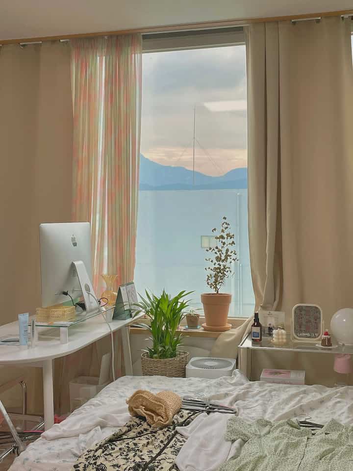 Beige and white toned studio bedroom featuring a window with sky view, curtains, computer desk, and plants in a cozy home office setting