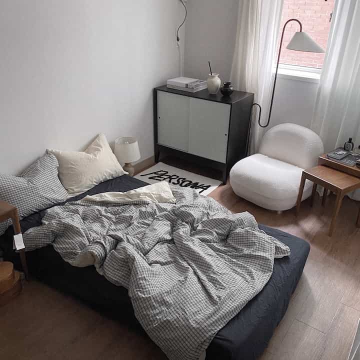 Natural and monotone studio bedroom featuring checkered bedding, a white armchair, and a black cabinet in a cozy space