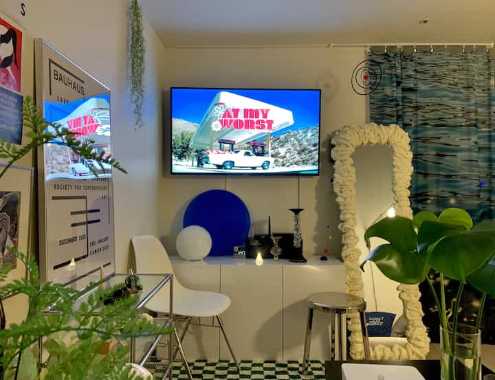 White and blue toned living room featuring mid-century modern poster, large TV, unique white textured mirror, and green plants creating a modern cozy atmosphere