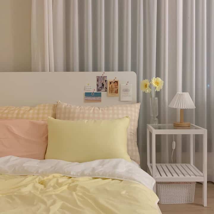 ホワイトとイエローのトーンの寝室、ベッドと枕、サイドテーブルの照明と花瓶が配置された落ち着いたシンプルな空間